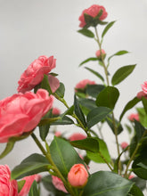 Charger l'image dans la galerie, This image captures a close-up of vibrant pink artificial flowers, with unopened buds and glossy green leaves, all set against a soft, blurred background. The composition highlights the delicate details of the petals and the natural arrangement of the leaves, giving the flowers a lifelike appearance.