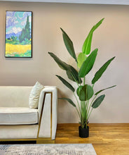 Load image into Gallery viewer, An elegant artificial bird of paradise plant with large green leaves is placed in a black planter on a hardwood floor beside a cream-colored modern sofa with gold accents. The room's beige walls create a warm backdrop, while a vibrant framed painting of a scenic landscape adds color and visual interest to the minimalist decor