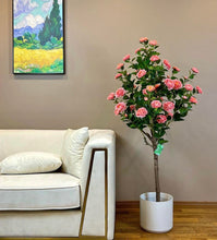 Charger l'image dans la galerie, This image shows a beautiful artificial pink flower arrangement placed in a white pot beside a cream-colored sofa. The flowers are lush and realistic, with dark green leaves adding to the natural effect. The background features a painting on the wall, adding an artistic touch to the cozy, elegant setting.