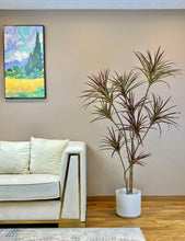 Charger l'image dans la galerie, Contemporary living room interior featuring a white sofa with gold accents and a faux Dracaena plant with red-tinted leaves in a white planter. The artificial plant complements the minimalist decor, alongside a vibrant wall painting, adding a touch of color and elegance to the space.