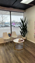 Load image into Gallery viewer, A modern waiting area in a commercial space featuring two sleek white chairs next to a large artificial bird of paradise plant. A round coffee table with a gold frame holds a tissue box and a small plate of treats. Large windows in the background offer a view of a parking lot with several cars, and the space is brightened by natural light.