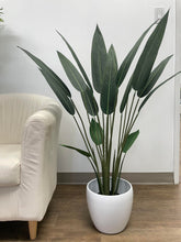 Load image into Gallery viewer, Artificial bird of paradise plant with long, slender green leaves, placed in a sleek white pot beside a beige armchair, creating a minimalist and modern indoor setting on a wooden floor.