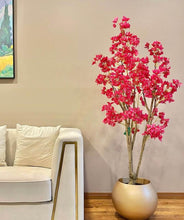 Charger l'image dans la galerie, A bright artificial flowering plant with pink blossoms placed in a gold round planter next to a beige sofa, set against a neutral-toned wall in a modern living room.