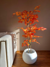 Load image into Gallery viewer, Artificial autumn maple tree with vibrant orange and red leaves in a white round pot, placed next to a modern white sofa against a warm-toned wall, with sunlight casting shadows on the floor