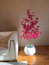 Load image into Gallery viewer, Artificial pink flowering tree in a white round planter, placed next to a beige sofa in a modern living room with wooden flooring.