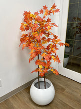 Load image into Gallery viewer, Artificial autumn maple tree with vibrant orange and red leaves in a white round pot, placed next to a modern white sofa against a warm-toned wall, with sunlight casting shadows on the floor