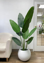 Load image into Gallery viewer, Tall artificial plant with large, broad green leaves in a round white planter placed beside a beige armchair, creating a modern and stylish indoor decor setup with a mirrored door in the background.