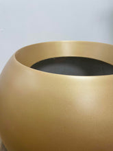 Charger l'image dans la galerie, Close-up view of a matte gold round planter with a smooth texture, placed on a wooden floor against a backdrop of green artificial leaves.