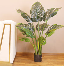 Charger l'image dans la galerie, Artificial monstera plant in a black pot, placed on a wooden floor next to a modern white and gold-trimmed armchair. The plant features large, glossy green leaves with distinctive splits, creating a tropical atmosphere in the indoor setting