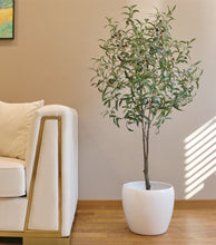 Load image into Gallery viewer, living room setup featuring a tall artificial olive tree in a white pot positioned next to a modern beige sofa with gold trim. The room has a warm, brown-toned wall adorned with a colorful landscape painting, creating a cozy and sophisticated atmosphere.