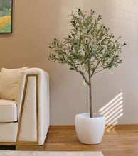 Load image into Gallery viewer, Elegant living room setup featuring a tall artificial olive tree in a white pot positioned next to a modern beige sofa with gold trim. The room has a warm, brown-toned wall adorned with a colorful landscape painting, creating a cozy and sophisticated atmosphere.