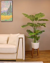 Load image into Gallery viewer, Artificial monstera plant in a white ceramic pot with a wooden stand, positioned next to a cream-colored armchair. The plant features large, realistic green leaves with natural splits, adding a tropical vibe to the indoor space. The setup is placed on a carpeted floor with subtle blue accents