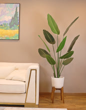 Load image into Gallery viewer, Artificial bird of paradise plant with tall green leaves in a white planter with wooden legs, placed next to a modern cream-colored sofa in a stylish living room with a framed landscape painting on the wall.