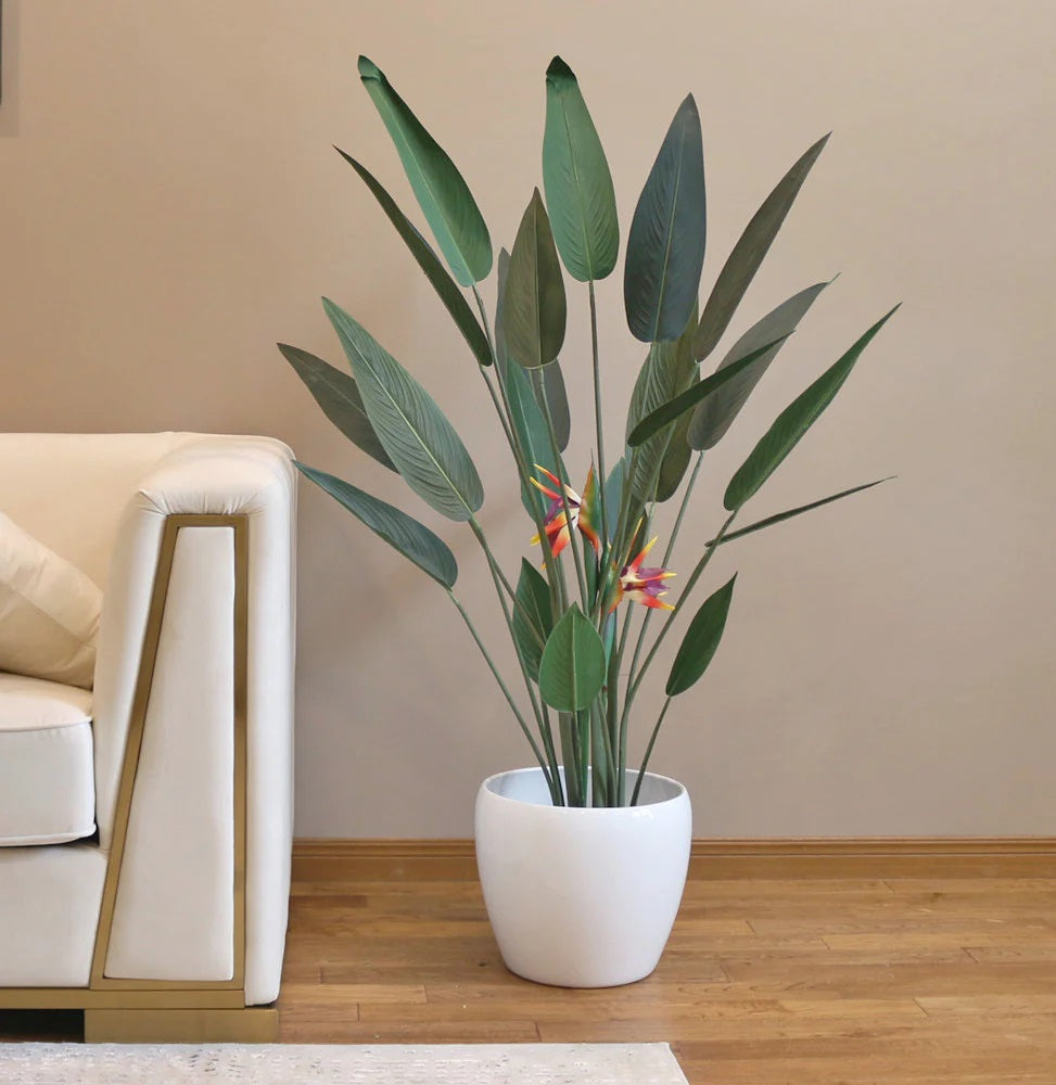 An artificial bird of paradise plant in a white pot placed in the living room besides a white sofa.