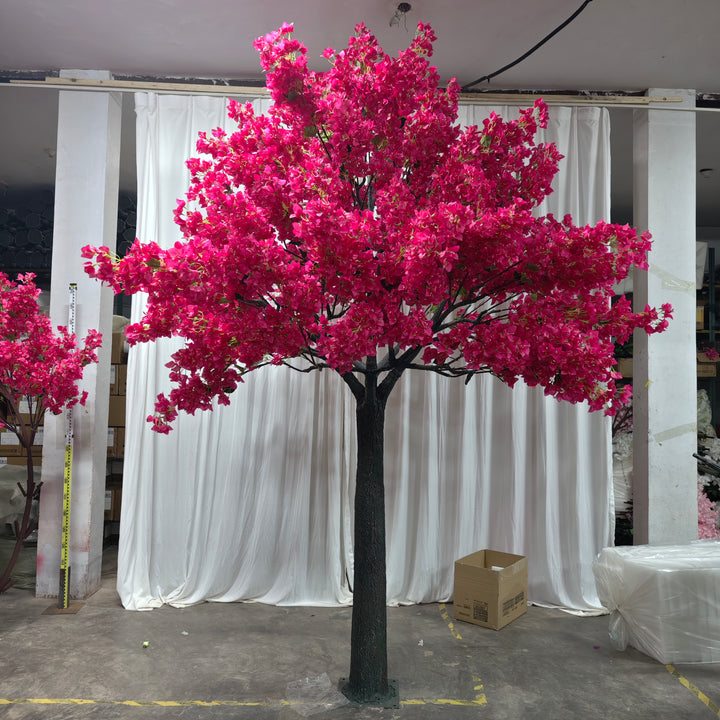A vibrant artificial bougainvillea tree with lush pink flowers on a dark green trunk, displayed indoors against a white draped backdrop. The scene includes cardboard boxes and work materials in the background, suggesting a workshop or preparation area.