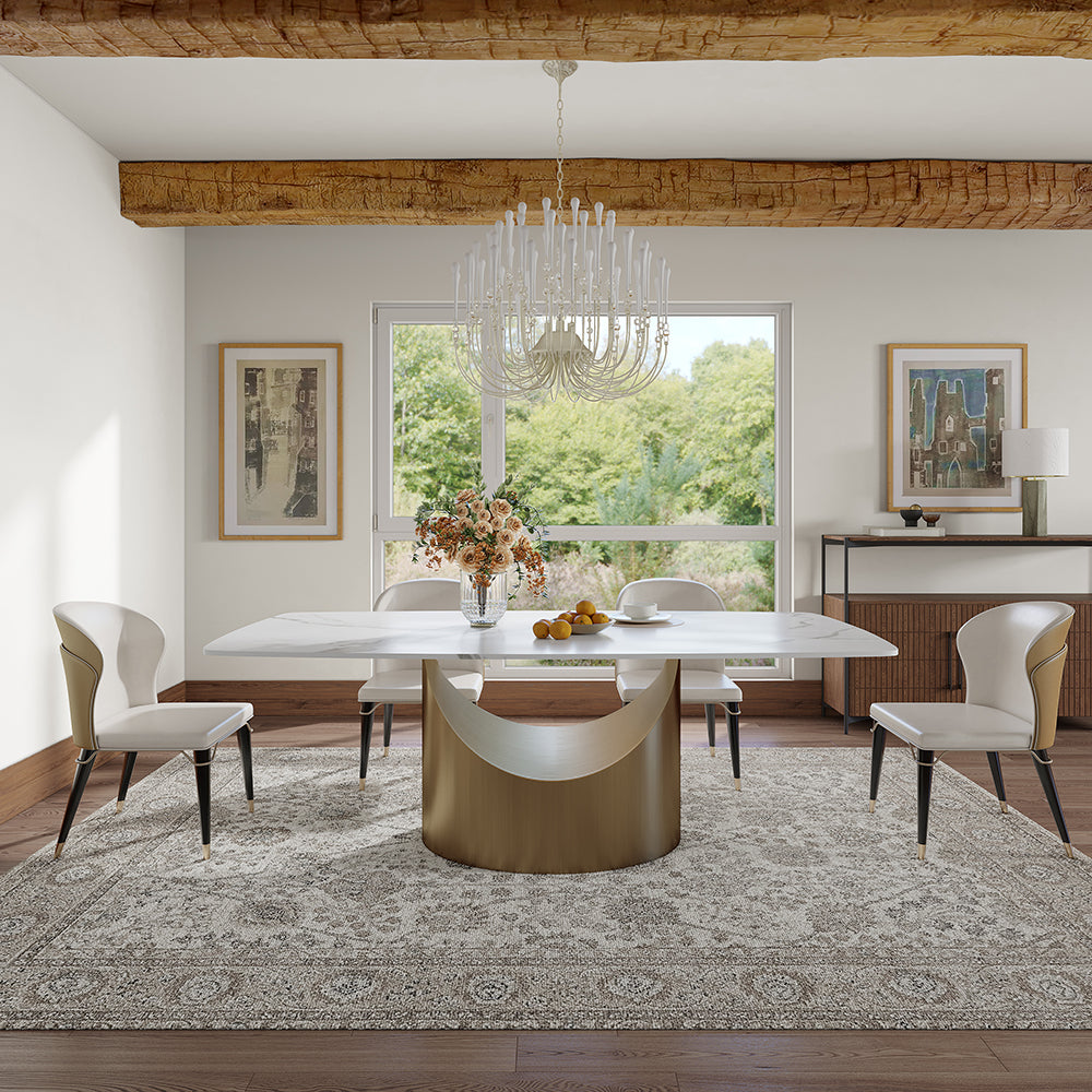 Rustic-chic dining room with a stunning oval marble table on a gold crescent base, paired with cream leather chairs. Crystal chandelier and exposed wooden beams add charm, while large windows frame a verdant garden view