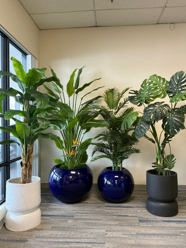 Four potted artificial tropical plants in various pots placed near a window.