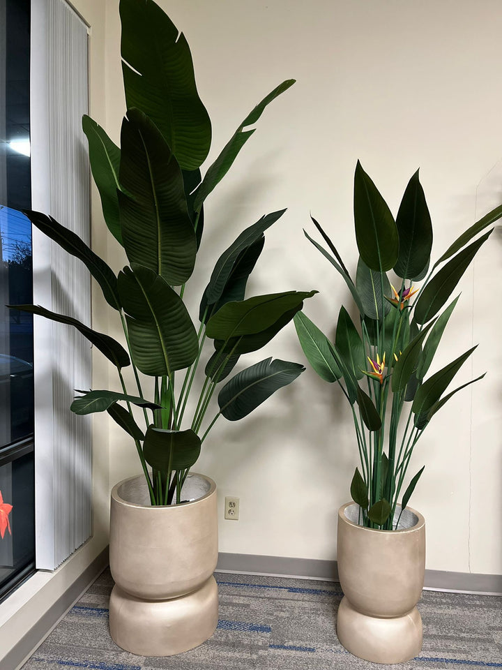 Pair of tall golden stackable planters with a smooth matte finish, each holding artificial plants with large green leaves, placed indoors near a window with natural light. Are these planters made from lightweight materials for easy mobility?"