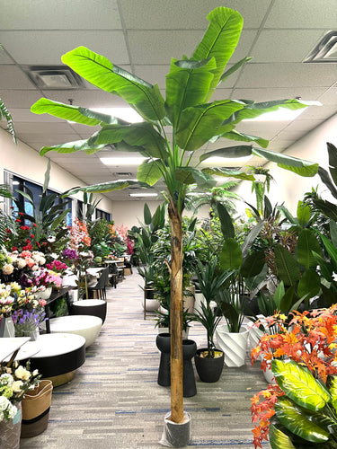 Tall artificial banana tree with large green leaves displayed in a well-decorated indoor showroom filled with various artificial plants and flowers