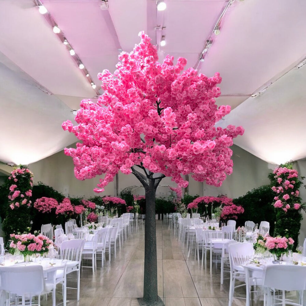A stunning artificial pink cherry blossom tree is prominently displayed in the center of an elegant event venue. The room features white chairs and tables adorned with pastel floral arrangements, set against a backdrop of lush greenery and a high, translucent ceiling that allows soft natural light to fill the space.