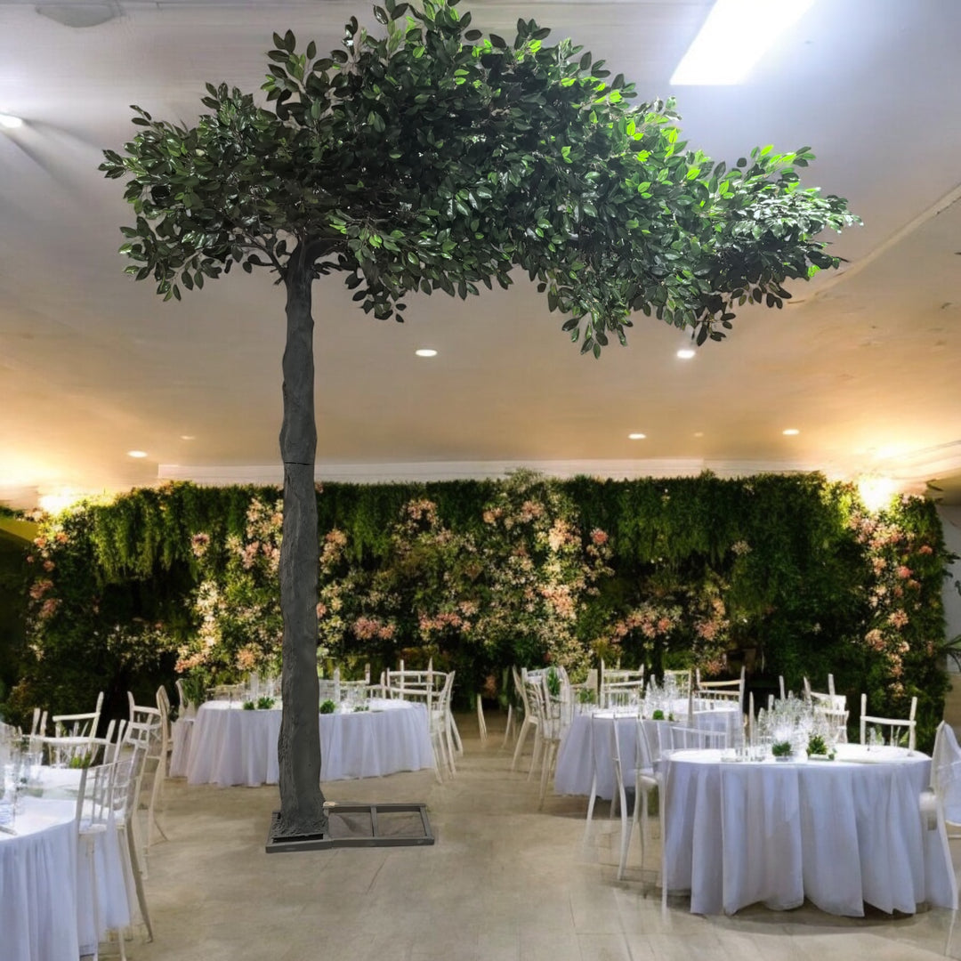Artificial green ficus tree with dense foliage, elegantly set in a wedding venue adorned with white round tables and a lush floral backdrop. 