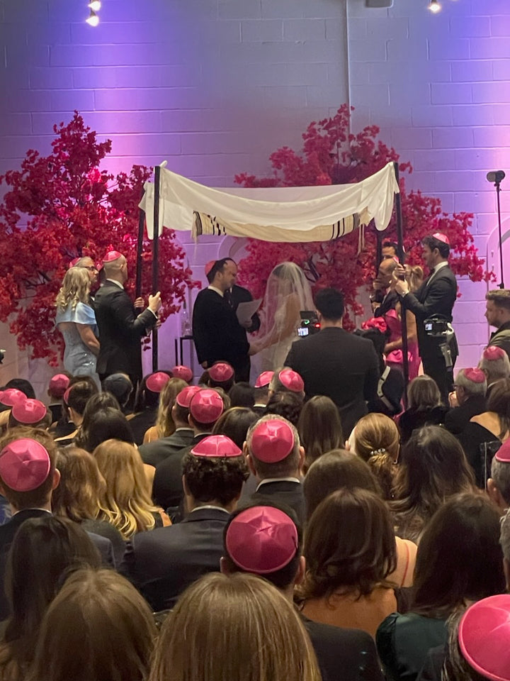 Wedding ceremony with hot pink bougainvillea  a chuppah, guests wearing pink yarmulkes, and a decorated background.