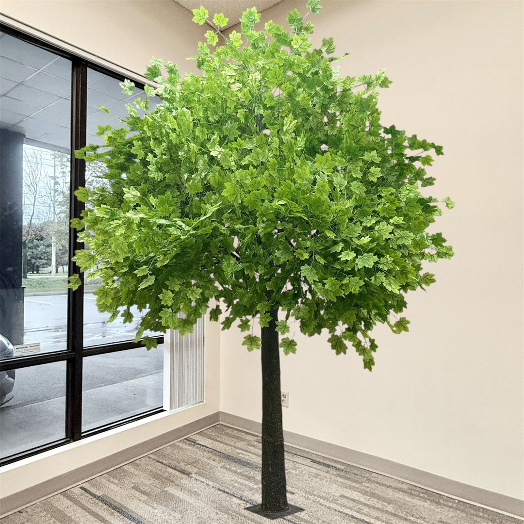 A large artificial maple tree with lush green leaves and a dark textured trunk, standing indoors near a window and beige wall.