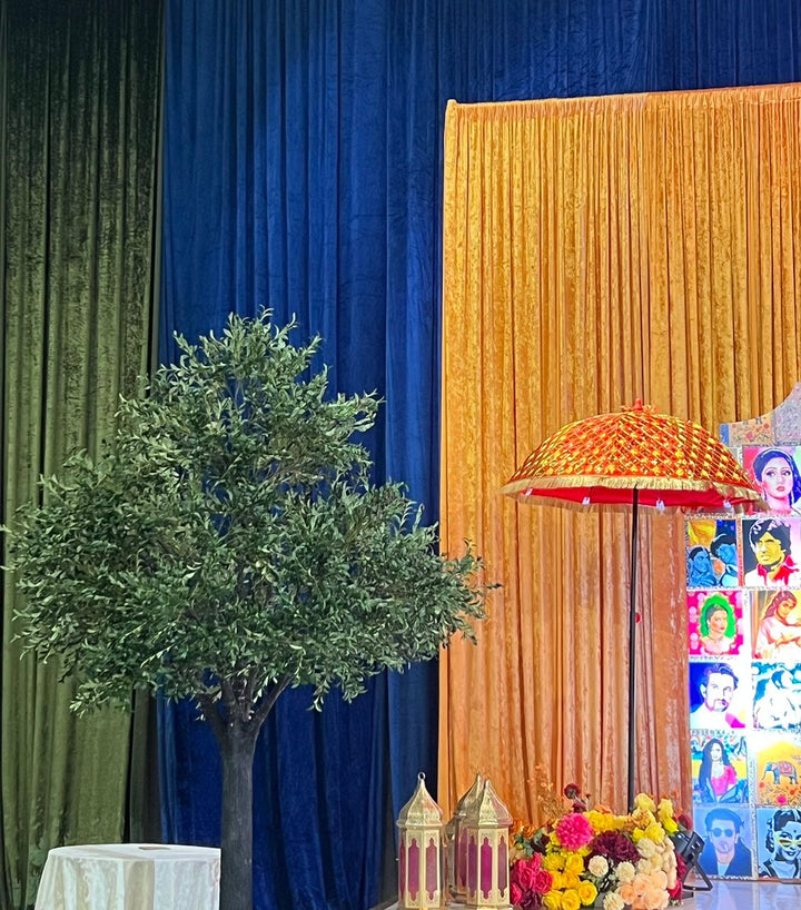 Decorative setup with green olive plant, colorful umbrella, and small buildings against a blue and gold curtain.