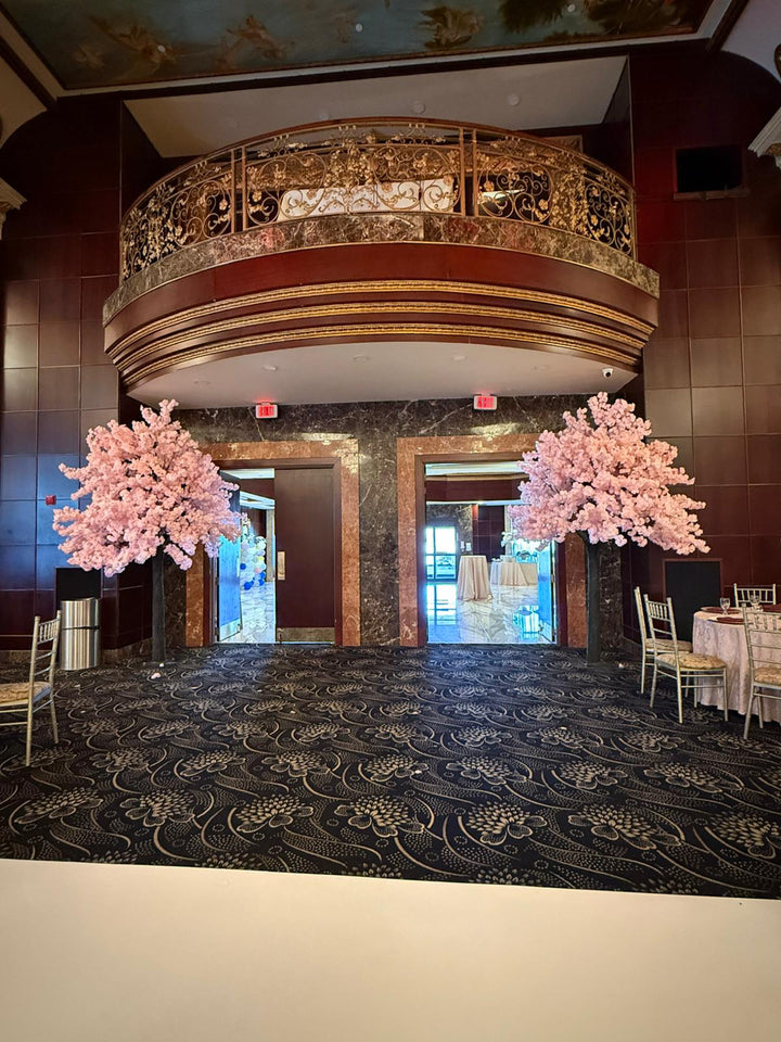 A large event hall with a patterned dark carpet, a decorative balcony with gold railings, and two pink cherry blossom tree decorations flanking double doors.