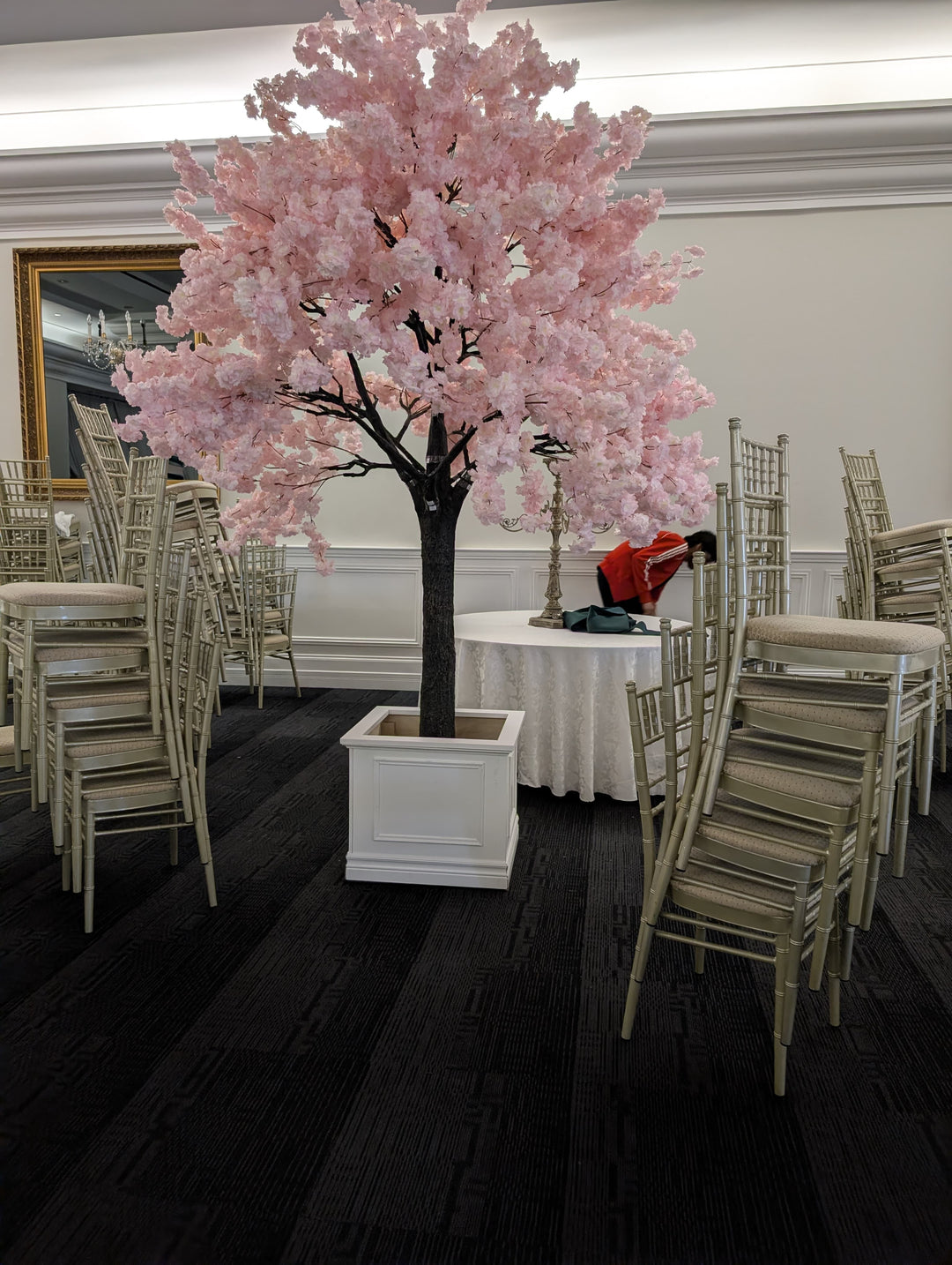 A large artificial pink cherry blossom tree in a white square planter displayed in a banquet hall or event space, surrounded by stacked champagne-colored chairs and a round white table.