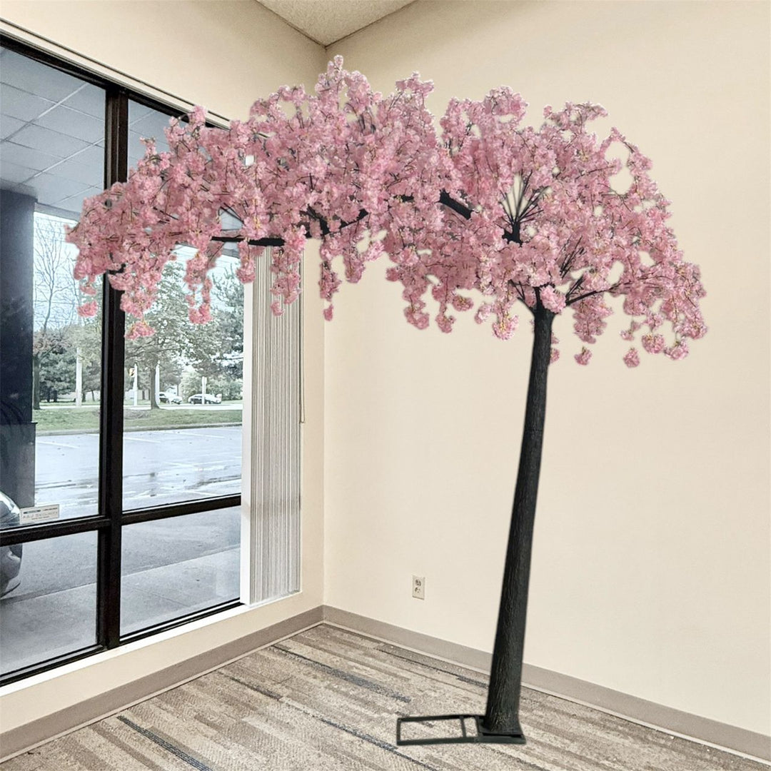 A large artificial cherry blossom tree with dense pink flowers and a dark trunk, displayed indoors in a room corner with a window, beige walls, and patterned carpet.