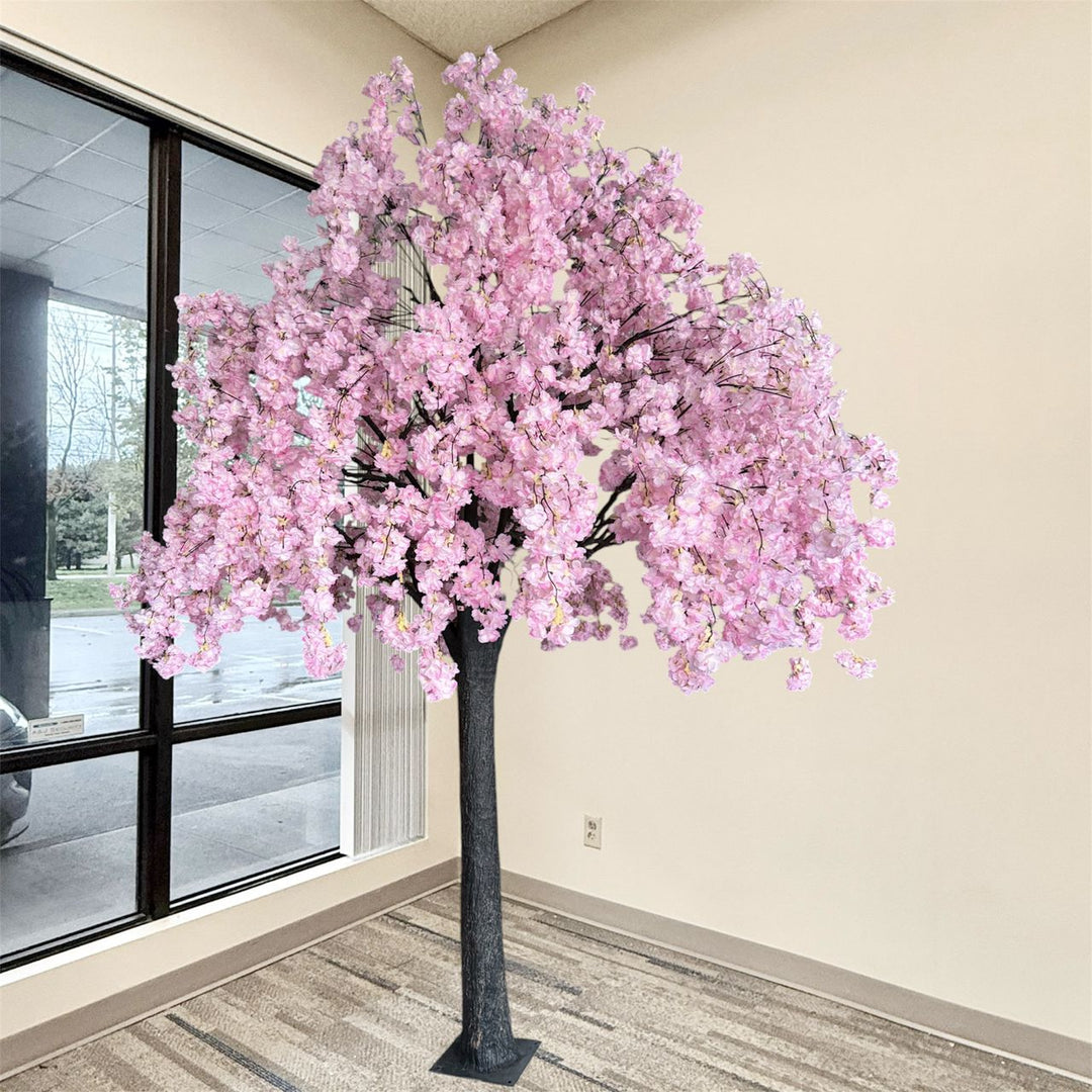 An artificial weeping cherry blossom tree with dense pink blossoms and a dark textured trunk, displayed indoors near a window and beige wall.