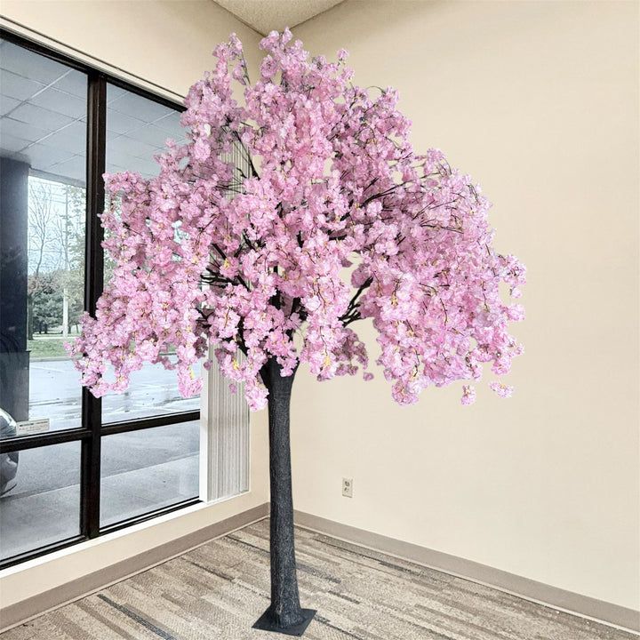 An artificial weeping cherry blossom tree with dense pink blossoms and a dark textured trunk, displayed indoors near a window and beige wall.