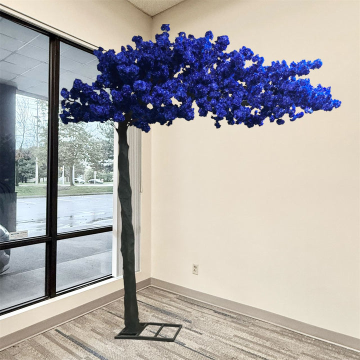 A large artificial cherry blossom tree with vibrant blue flowers and a dark textured trunk, displayed indoors in a room with beige walls and a window.
