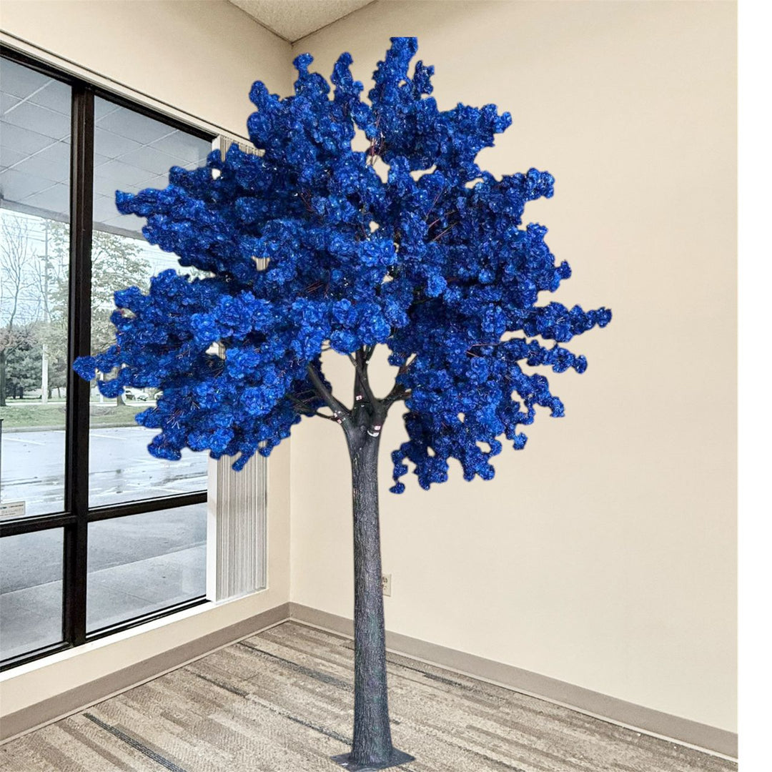 A decorative artificial tree with dense, vibrant royal blue foliage and a textured gray trunk, standing in a room corner with beige walls and a window.