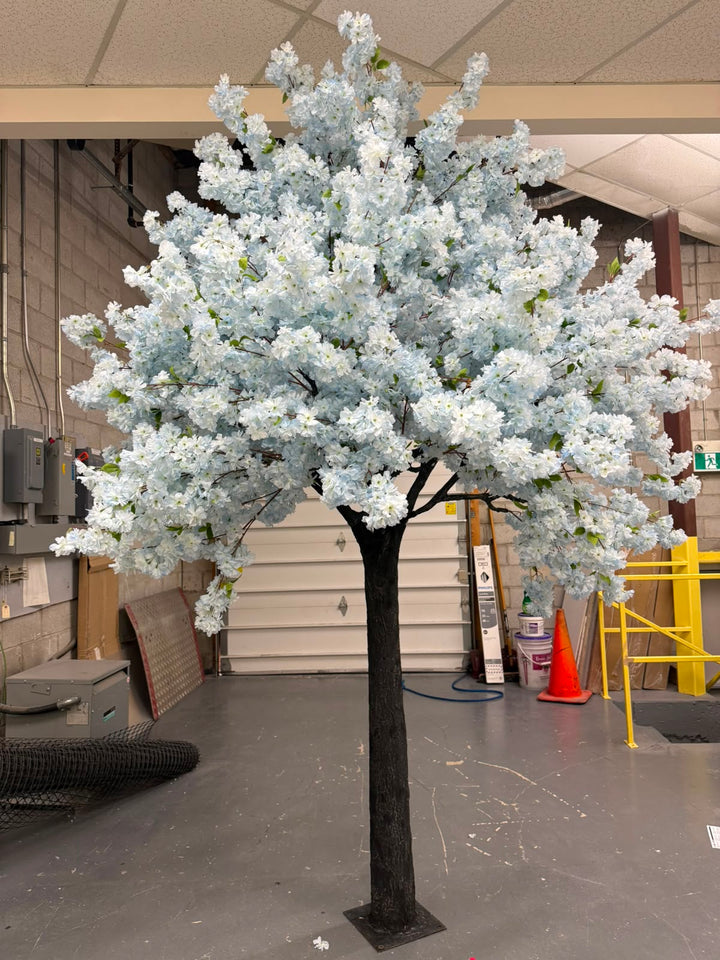 An artificial flowering tree with  soft  blue blossoms and a dark trunk, standing indoors in a workshop or warehouse setting.