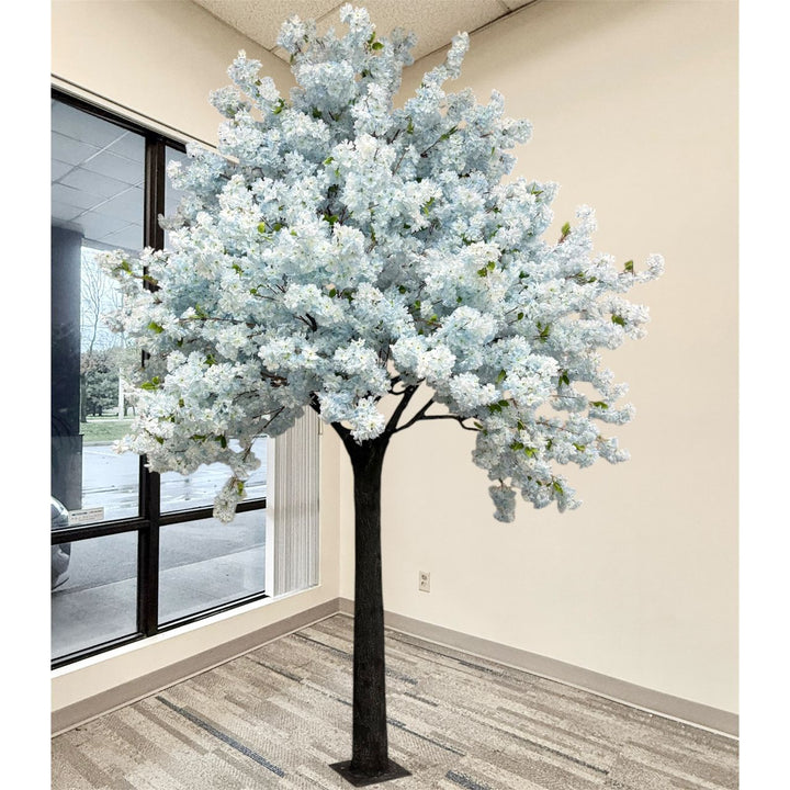 An artificial  soft blue cherry blossom tree with dense soft blue flowers and a dark trunk, displayed indoors in a room with beige walls and a window.