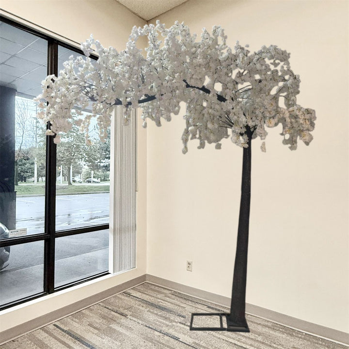 A large artificial weeping cherry blossom arch tree with white flowers and a black trunk, displayed indoors in a room with beige walls and a window.