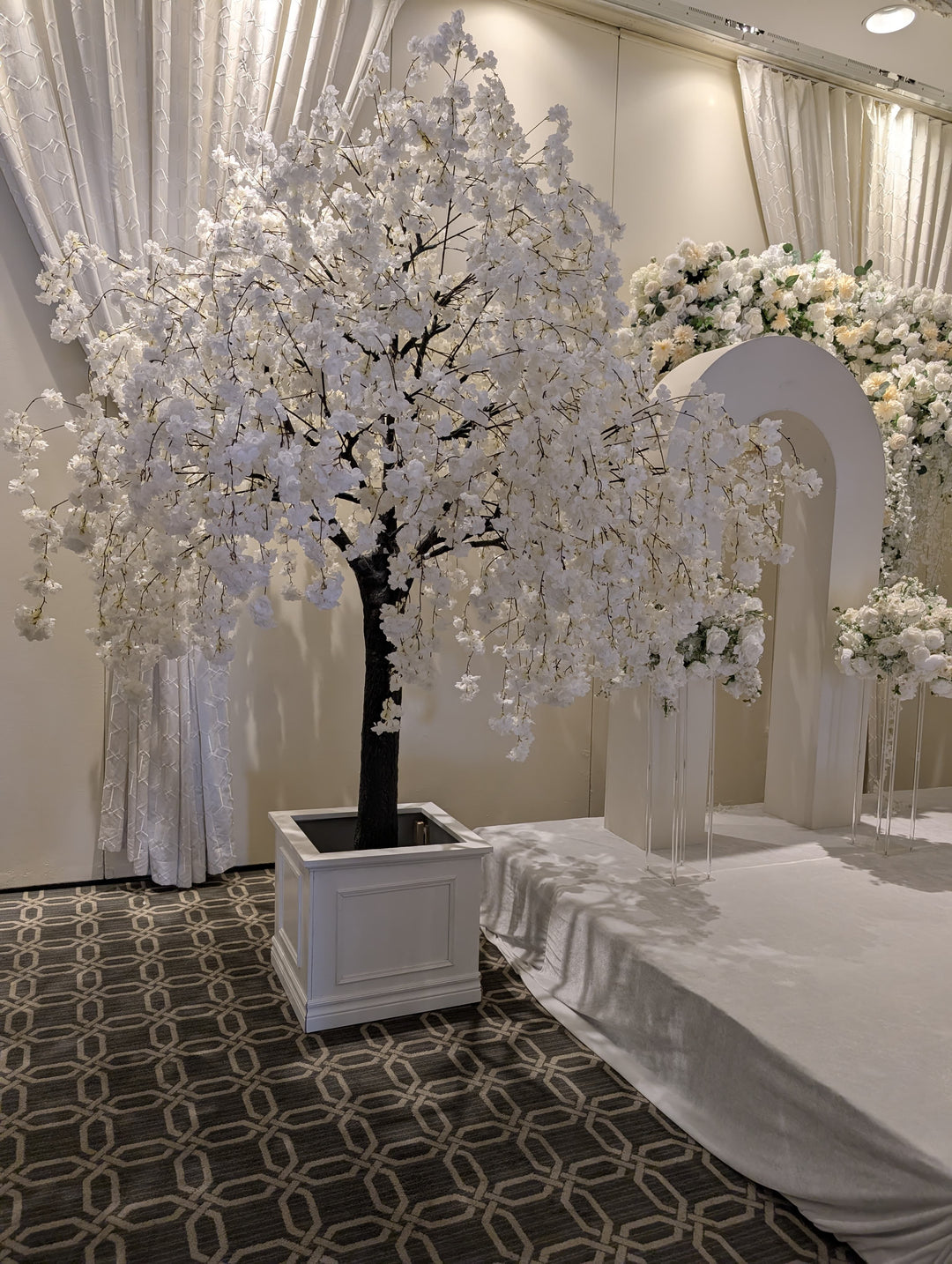 Decorative white floral  weeping cherry blossom tree in a pot on a patterned carpet with draped curtains in the background in Toronto.