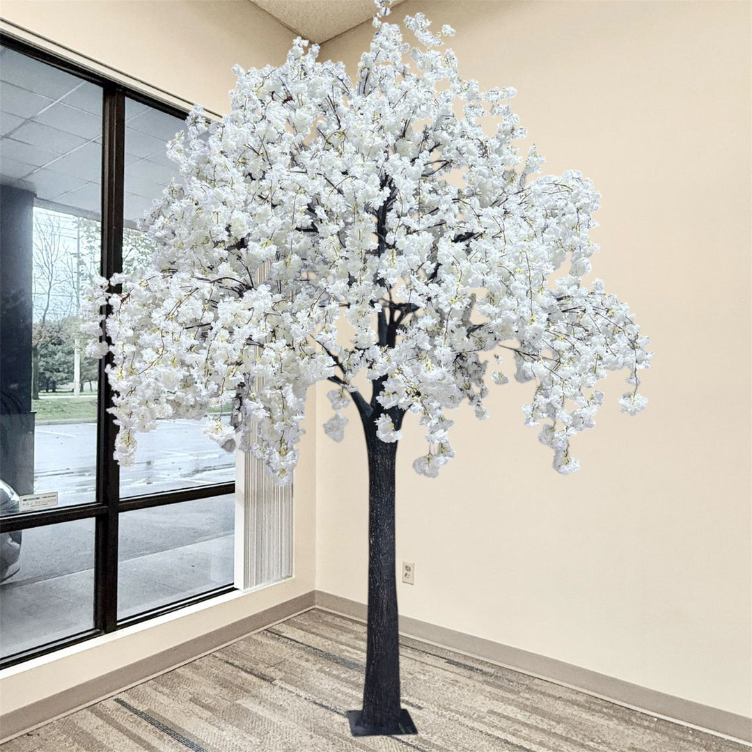 An artificial  white weeping  cherry blossom flowering tree with dense white blossoms and a dark textured trunk, standing indoors near a window and beige wall.
