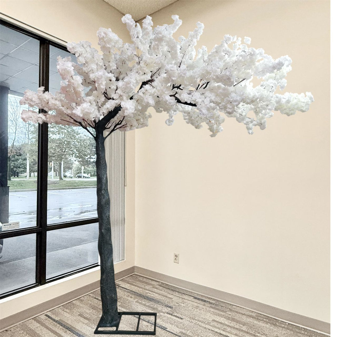 A large artificial cherry blossom tree with white faux flowers and a dark textured trunk, displayed indoors near a window in a beige room.