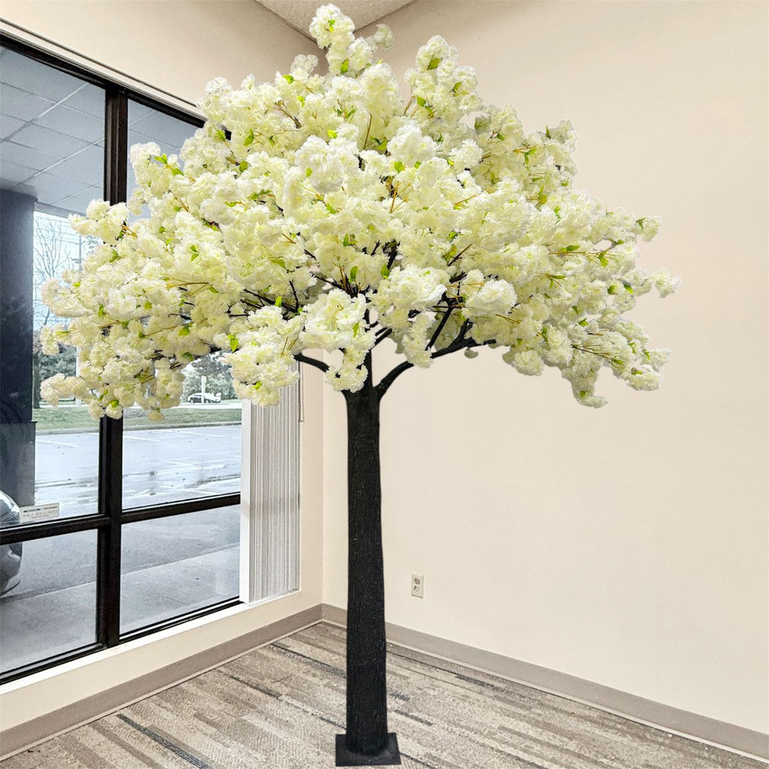 An artificial cream white flowering tree with dense white blossoms and green leaves  and a black trunk, standing indoors near a large window with a beige wall and wood-look floor.