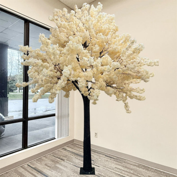 An artificial  cream white flowering cherry blossom tree with dense cream-white blossoms and a black trunk, displayed indoors against a beige wall next to a large window.