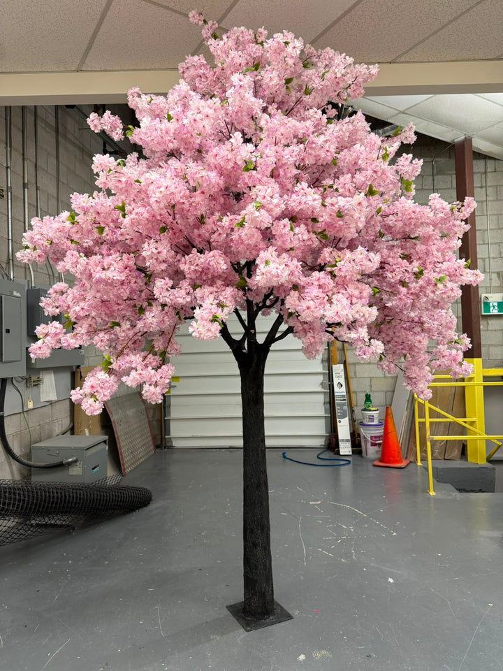 An artificial light purple cherry blossom tree with dense white pink flowers and a dark trunk, standing on a gray floor inside a warehouse or workshop.