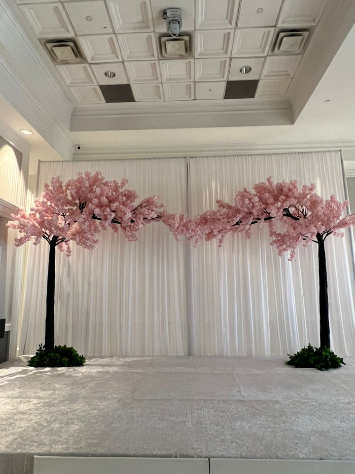 A decorative event backdrop setup featuring two artificial pink cherry blossom trees flanking a white draped curtain, with a light gray carpeted stage in front.