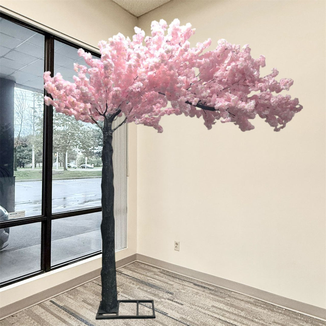 A large artificial light  pink arch cherry blossom tree with soft pink blossoms and a dark textured trunk, set indoors next to a window with beige walls and striped carpet.