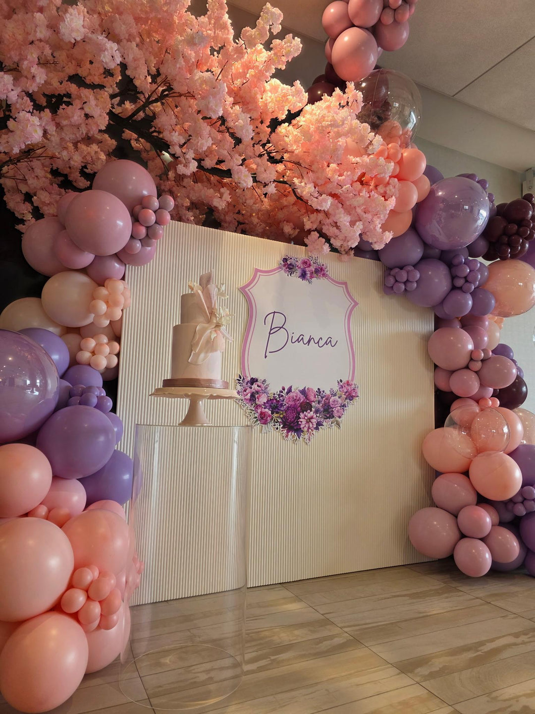 Decorative setup with balloons, arch peach cherry blossom flowers, and a cake display in a room.
