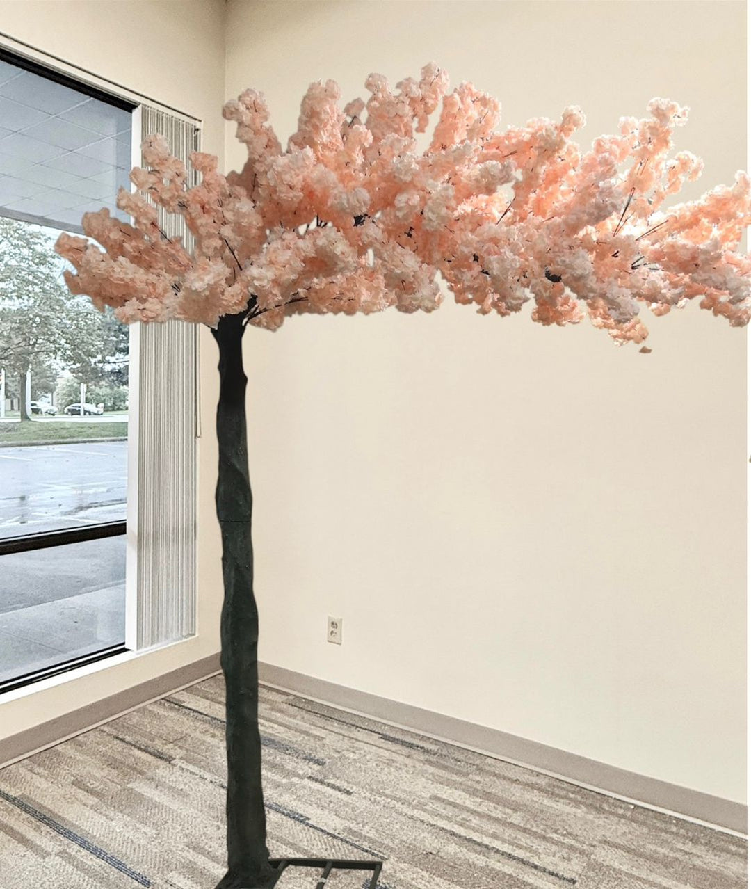 An artificial cherry blossom tree with peach blossoms and a dark textured trunk, standing indoors near a window with a gray patterned floor.
