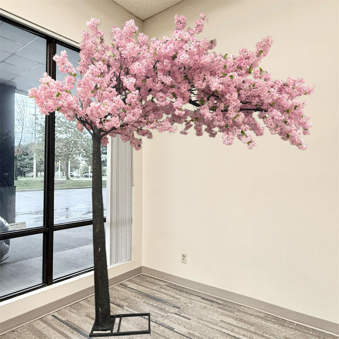 An artificial  purple cherry blossom tree with dense purple blossoms and a dark trunk, displayed indoors in a room with beige walls, a window, and a patterned carpet.