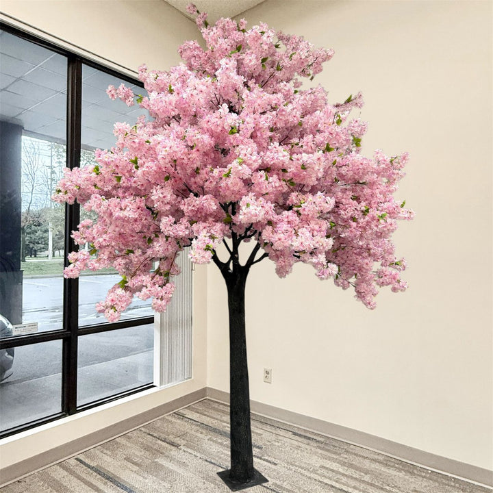 A large artificial cherry blossom tree with dense purple flowers and a dark trunk, displayed indoors near a window in a room with beige walls and gray-brown flooring.